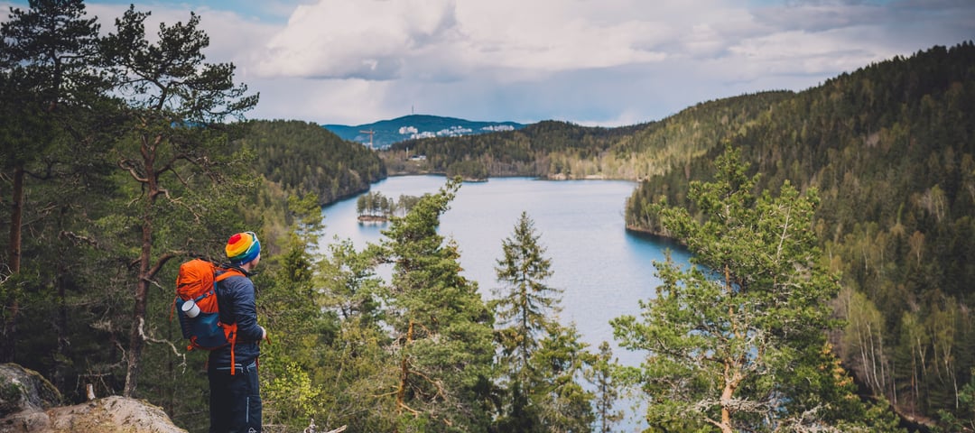 mann med sekk og regnbuelue som står på en høyde og ser utover et vann og skog