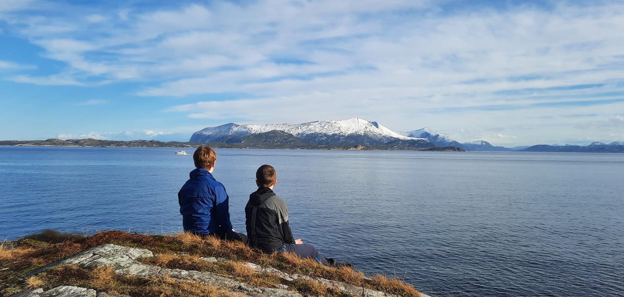 To personer sitter på berget på Sandvikstien i Kristiansund med ryggen til. De har utsikt mot Tustnafjella