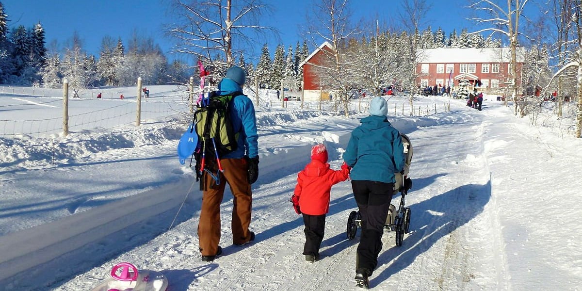 To voksne og et barn triller en barnevogn opp bakken mot en rød gårdsbygning om vinteren, med snødekte bakker rundt. Masse folk er samlet rundt hytta, og noen leker i snøen. 