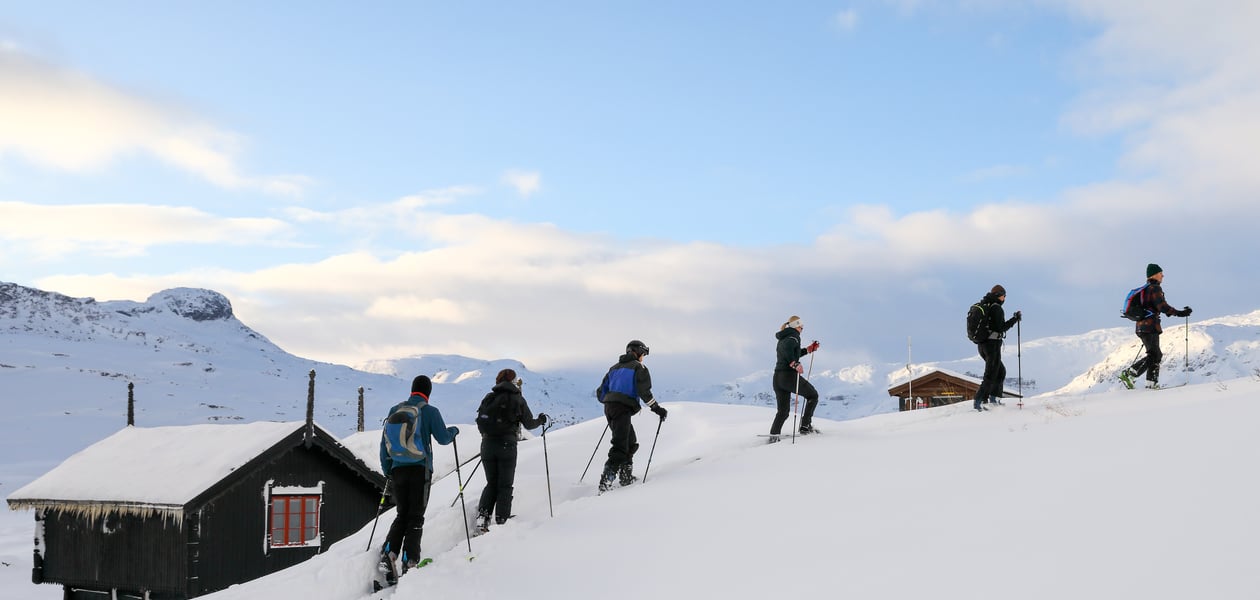 Foto. En rekke med skigåere sett fra siden. Blå himmel, snø og et laftet stabbur i bakgrunnen.