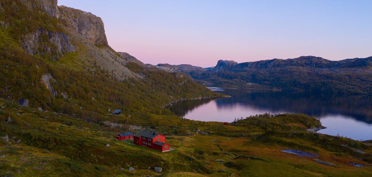 En rød hytte i solnedgang som ligger i et storslått fjellterreng med et vann i sentrum av fjellene.