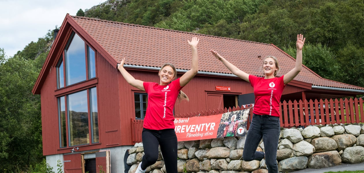 To jenter i røde t-skjorter hopper på plenen utenfor en rød låve.
