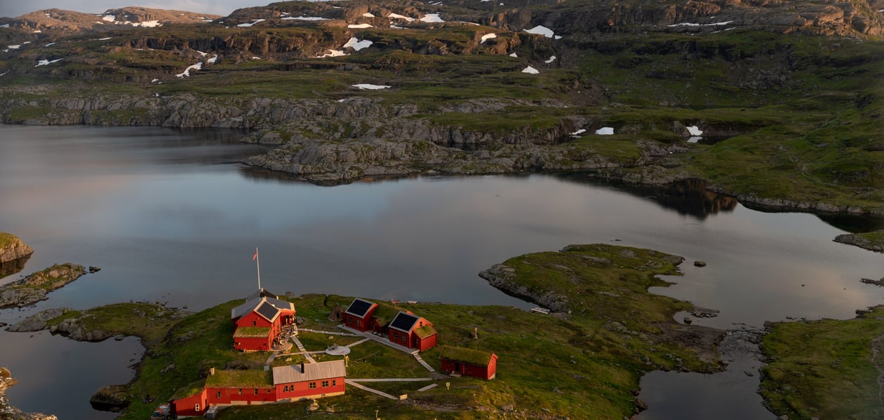Dronebilde av hytta med oversikt over hytta, vannet og fjellene.