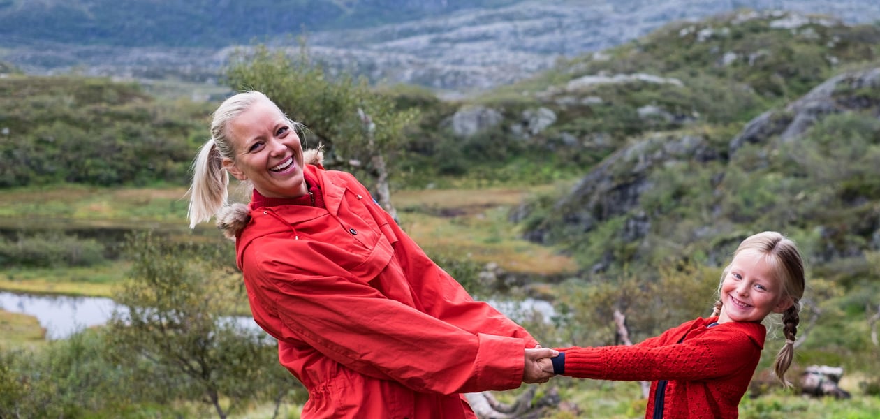 Mor og datter i rødfargede klær holder hender, lener seg tilbake og smiler mot kamera. Landskapet er grønt og på fjellet.