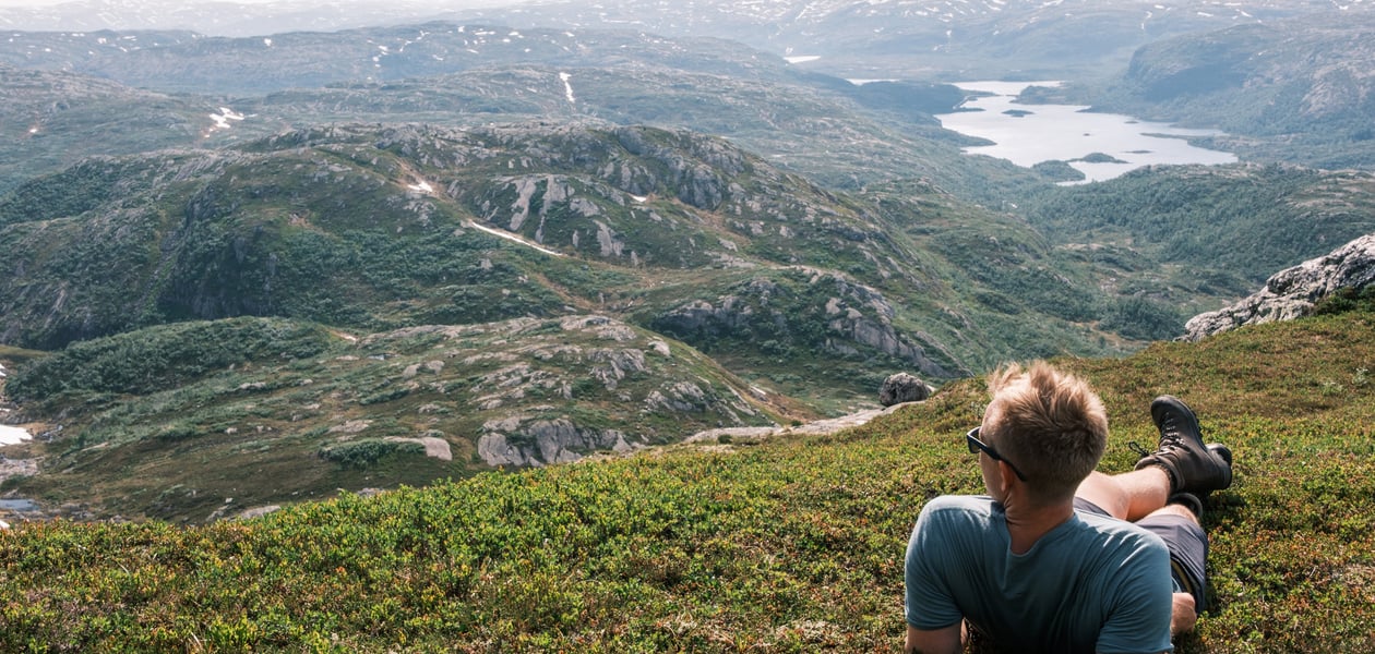 En mann som har solbriller, shorts, t-skjorte og fjellsko, ligger i gresset og ser utover fjellene