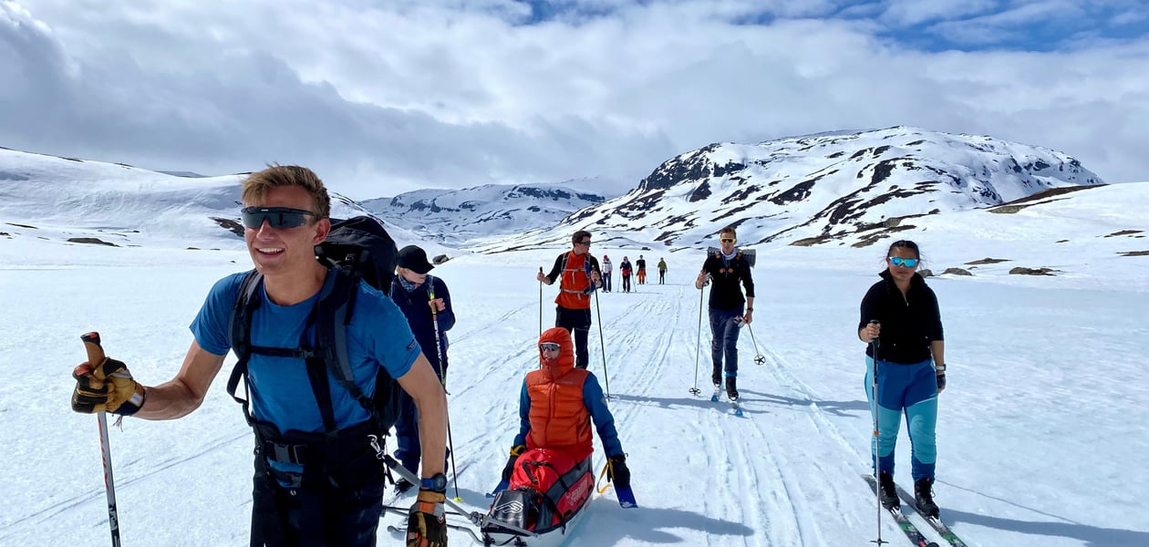 En mann på ski som har en annen handikappet mann i pulk. Flere i følget bak. Mannen i front smiler godt.