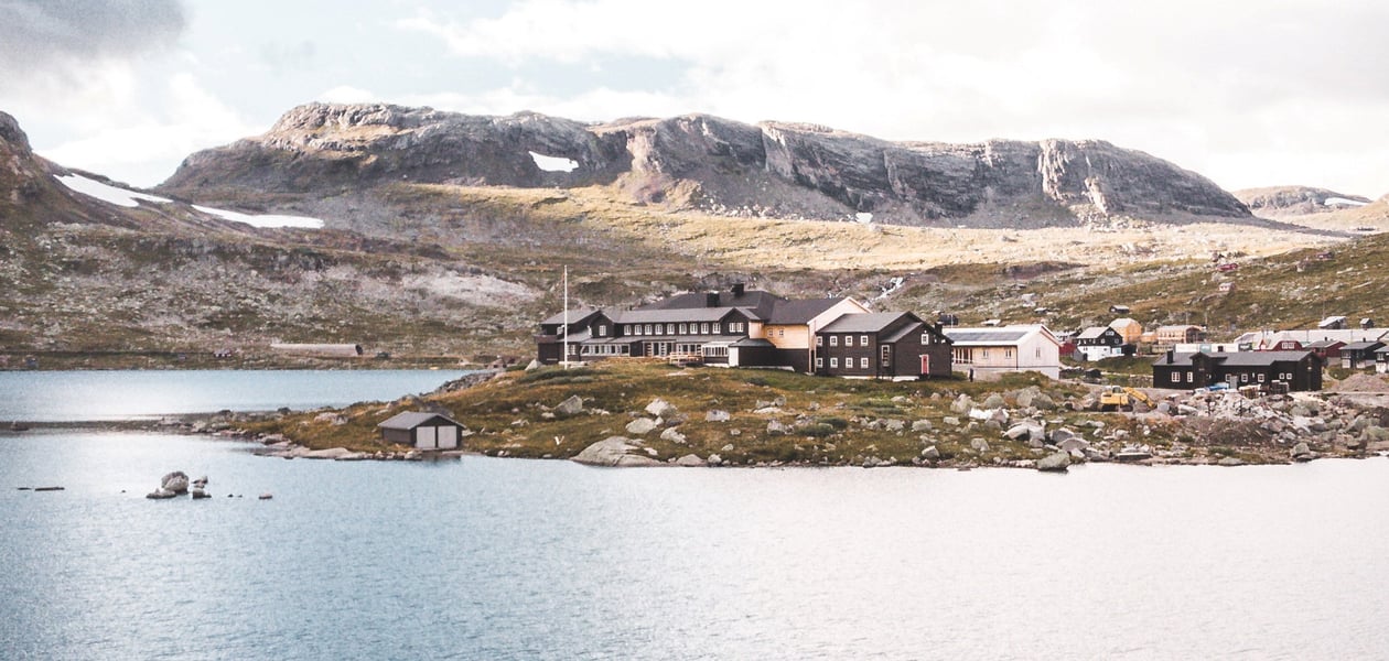 Dronebilde av en svartmalt hytte som ligger ved en innsjø og med fjell i bakgrunnen