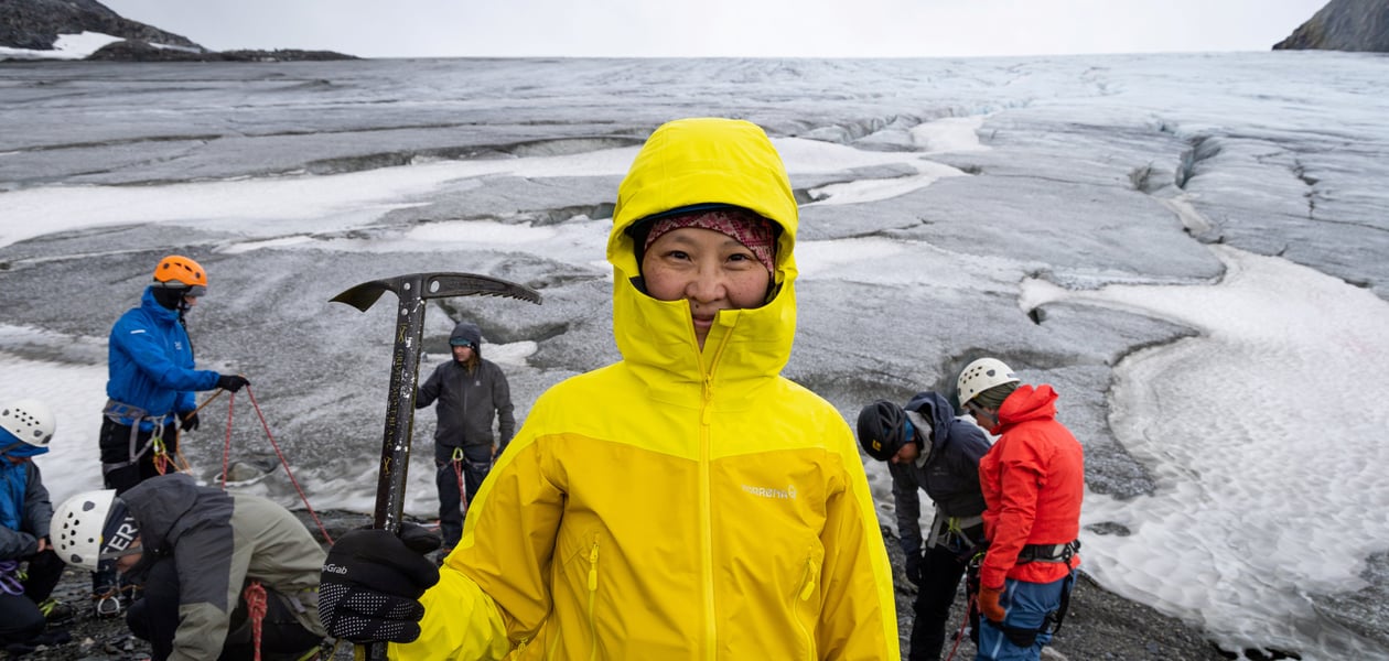 Mongolsk kvinne i gul jakke og isøks i hånden smiler i kamera foran isbre.