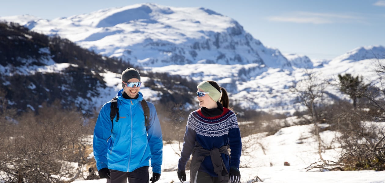 Mann i blå jakke og kvinne i mariusgenser smiler og går på tur i fjellet med snø og lyng.
