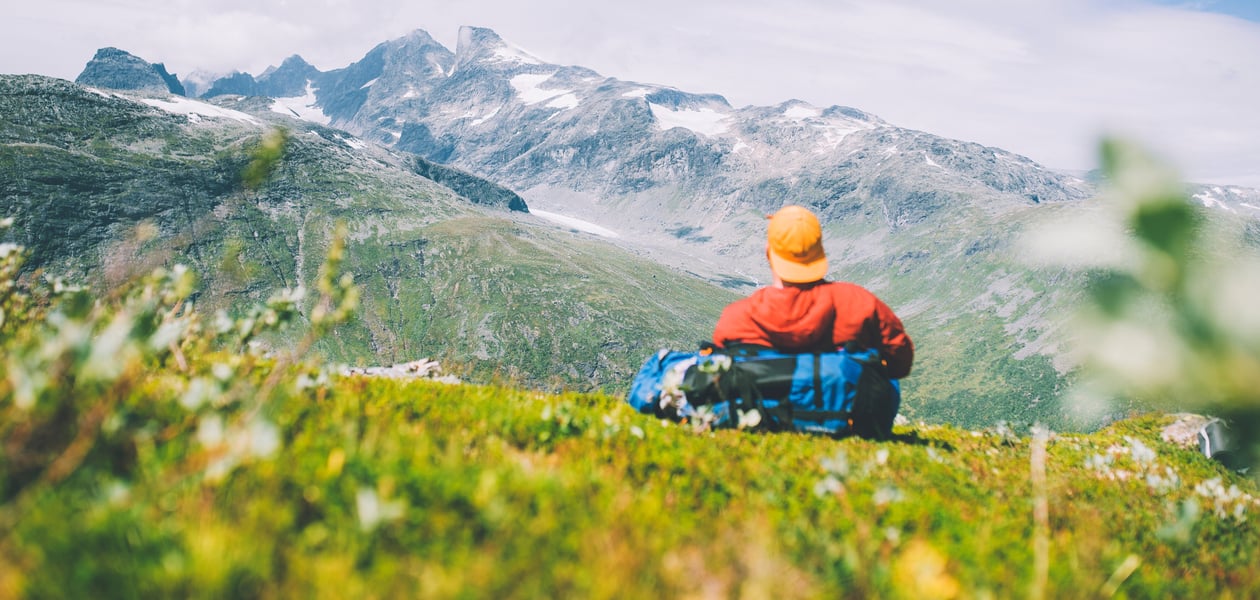 En mann i rød jakke og gul caps som sitter i lyngen og ser på fjell