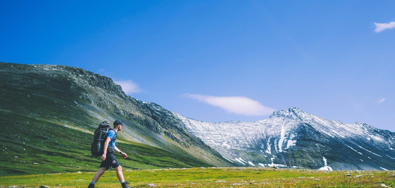 En person som går over en gresslette med stor sekk på ryggen. Store fjell med snø på toppene i bakgrunnen.