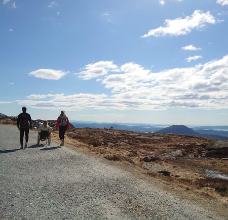 To gående og en trillende på tur på grusvei, med blå himmel og fjell i bakgrunnen