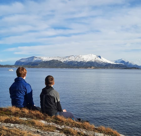 To personer sitter på berget på Sandvikstien i Kristiansund med ryggen til. De har utsikt mot Tustnafjella