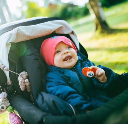 En baby med rosa lue og blå jakke sitter i en barnevogn utendørs, smiler og holder i en smokk, med sollys i bakgrunnen.