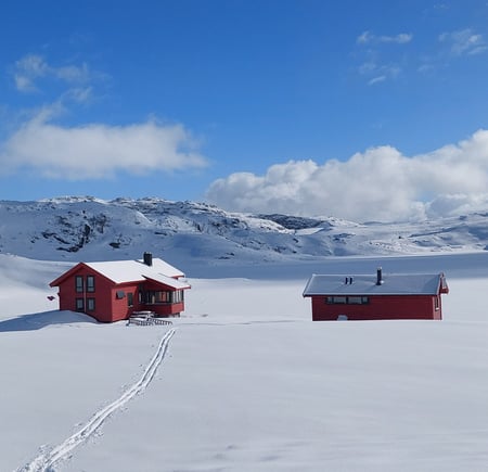 Tomannsbu. 2 røde hytter i snødekt landskap. Det er blå himmel og sol. 