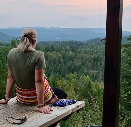 Jente sitter på en benk utenfor Tyristua i Botnemarka og ser på solnedgangen med utsikt til skog og åser.