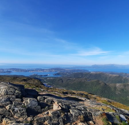 Bildet er tatt fra det som ser ut som en fjelltopp, med utsikt over skog og hav. Det er blå himmel.