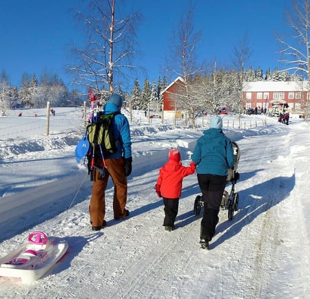To voksne og et barn triller en barnevogn opp bakken mot en rød gårdsbygning om vinteren, med snødekte bakker rundt. Masse folk er samlet rundt hytta, og noen leker i snøen.