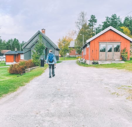 En person med ryggsekk går på en grussti mellom trebygninger, deriblant en rød hytte, i et landlig område med trær og gress.