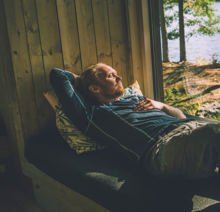 En person hviler på en sofa ved et vindu som vender ut mot en skog og en innsjø. Solstråler skinner gjennom trærne og skaper en fredelig atmosfære. Personen ser avslappet ut og nyter den rolige omgivelsen inne i en hytte
