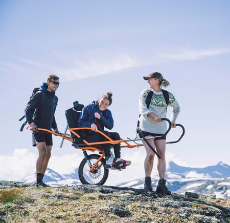 To personer transporterer en tredje person i rullestol med store hjul i fjellet. 