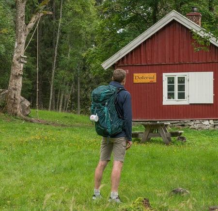 Person med sekk foran en rød hytte. 