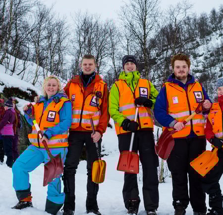 Fem personer med oransje vester og hver sin spade står i snøen og smiler