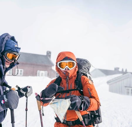 To skiløpere i vinterfjellet, i sterk vind, med hette, briller og kart.