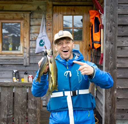 Mann i blå jakke smiler foran hytte og peker på noen fjellørret han har fanget.