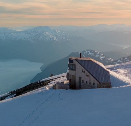 Langstrakt hytte på toppen av fjell i solnedgang med fjord lenger bak.