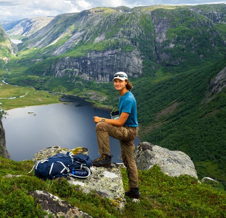 En mann som står på toppen av et fjell med grønt landskap rundt og et vann nederst i bildet.