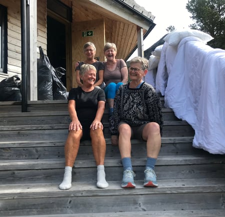 Fire damer sitter på en trapp utenfor en grå hytte. Det henger dyner til lufting.