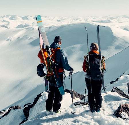 To personer med sekker hvor det er festet på ski på. De står og ser utover et snødekket landskap