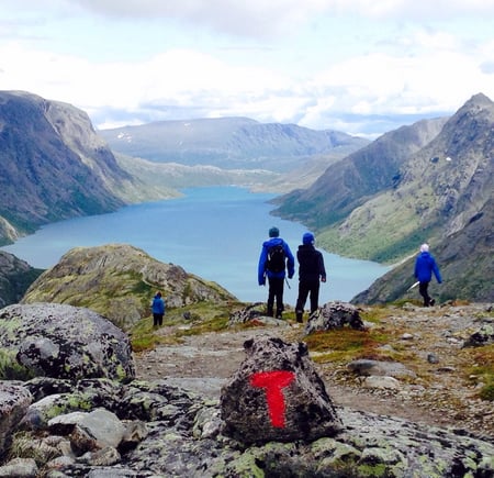 Personer og T-merket stein på fjelltopp, med utsikt mot vann og fjell i bakgrunnen