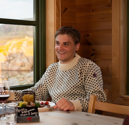 En mann sitter ved et bord og smiler. Foran han er det en tallerken med mat og et norsk flagg.