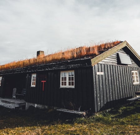 Svartmalt hytte med torvtak og en stor t på veggen. 