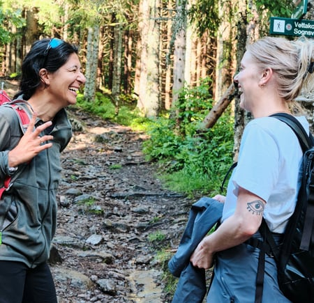 To personer smilende og pratsomme på en skogsti i Oslomarka. Begge bærer tursekker, og i bakgrunnen sees et skilt som viser retningene til Vettakollen (1,6 km) og Sognsvann (3,6 km)