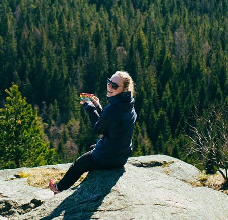 En kvinne sitter på en stein ute i naturen med en skogkledd åsside i bakgrunnen. Hun smiler mens hun holder en Kvikk Lunsj-sjokolade i hånden og har på seg solbriller og en mørk turjakke. Det ser ut som hun nyter en pause på en tur i skogen