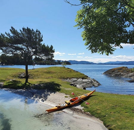 Kajakk ligger på sandstrand i idyllisk frilkuftsområde med sol, sjø og grønn gressplen.