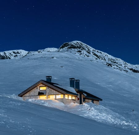 Hytte i vakkert snølandskap i fjellet med lys inne og stjernehimmel over hytta.