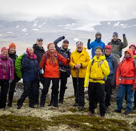 En turgruppe smiler og hoier på fjelltopp med skoddekledde fjell i bakgrunnen på høyfjellet.