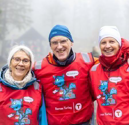 Tre frivillige med røde vester fra Turistforeningen smiler til kamera, en mann i midten som holder rundt to damer.