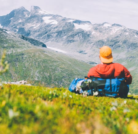 En mann i rød jakke og gul caps som sitter i lyngen og ser på fjell