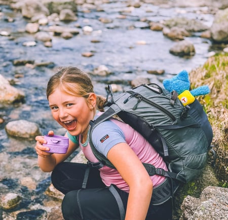 En liten jente sitter på huk og drikker fra en lilla kopp ved en elv. Hun smiler til kamera.