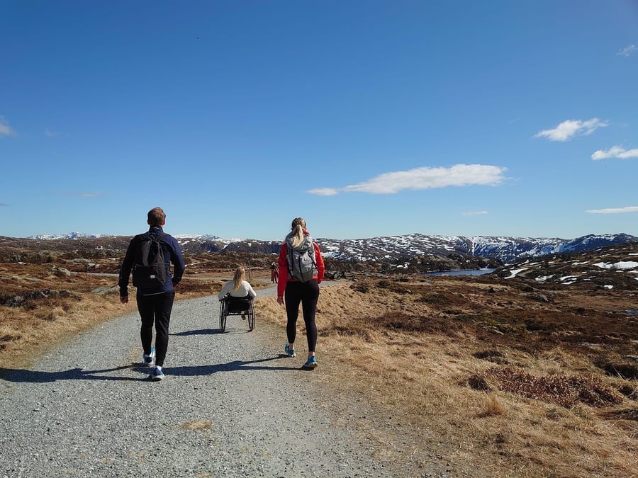 To gående og en trillende er på tur på grusvei, med blå himmel og fjell i bakgrunnen