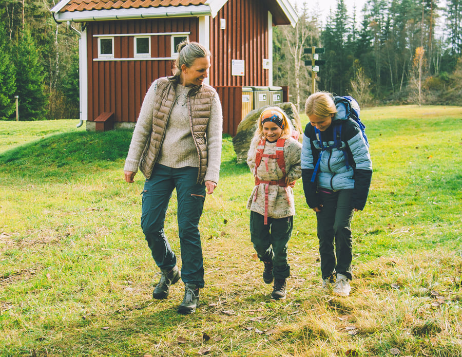 En voksen og to barn, alle kledd i friluftsklær, går sammen på en gresslette i nærheten av en rød trebygning med trær i bakgrunnen.