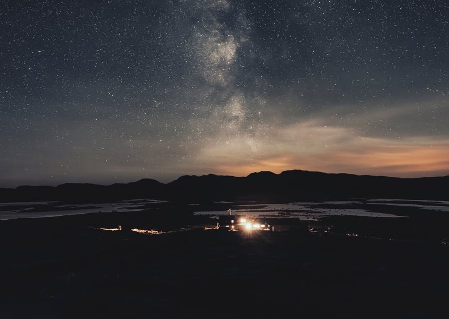 Et fjellandskap om natten med en klar stjernehimmel, hvor Melkeveien er synlig over horisonten, og et svakt opplyst bygg ses i det fjerne.