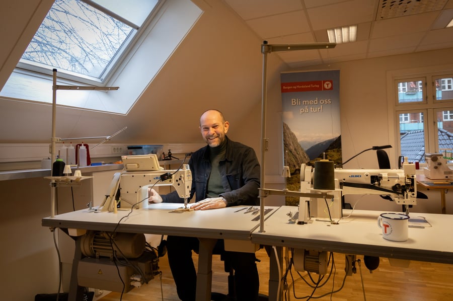 Mann ved symaskin i loftstue smiler til kamera.