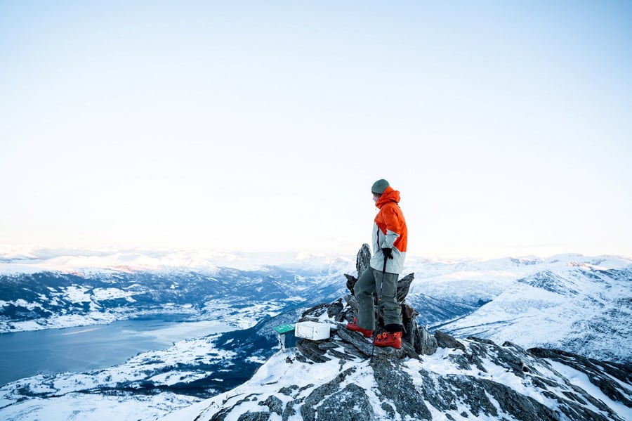person i vinterklær og slalåmstøvler, står på en delvis snødekket fjelltipp og ser utover et fjell- og fjordlandskap.