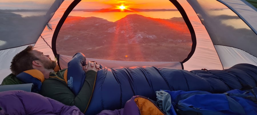 Bilde av innsiden av et telt, hvor en mann ligger i sovepose og ser ut gjennom bakveggen av teltet, hvor en solnedgang avgir sterkt oransje lys.